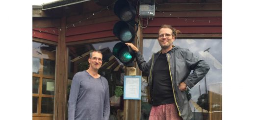 Zwei Männer stehen vor einem Gebäude, an dem die Energie-Ampel hängt. Sie leuchtet grün, was bedeutet, dass im Ökodorf Sieben Linden mehr Strom produziert wird, als aktuell gebraucht wird.