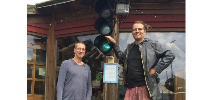 Zwei Männer stehen vor einem Gebäude, an dem die Energie-Ampel hängt. Sie leuchtet grün, was bedeutet, dass im Ökodorf Sieben Linden mehr Strom produziert wird, als aktuell gebraucht wird.