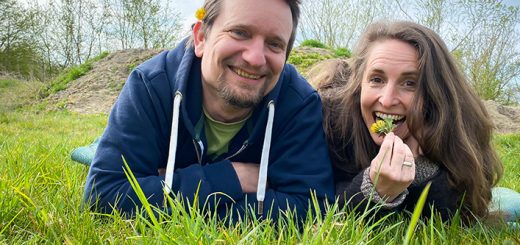Simone und Jörg liegen nebeneinander auf einer Wiese. Simone beißt in eine Löwenzahlblüte. Jörg lächelt in die Kamera.