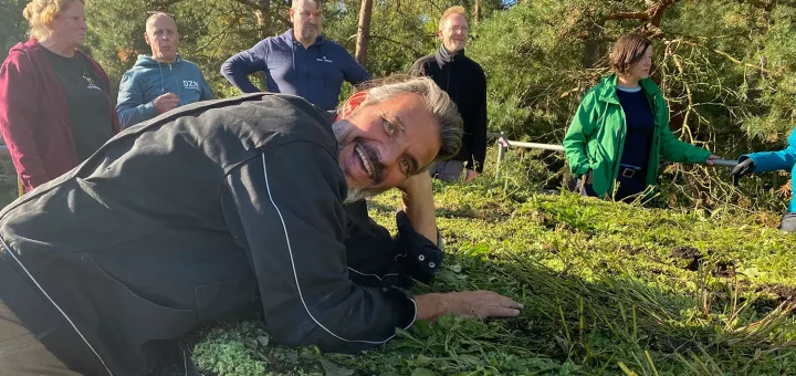 Rolando stützt sich auf ein Gründach und lächelt in die Kamera. Im Hintergrund steht die Seminargruppe, die bei der Begrünung des Dachs geholfen hat.